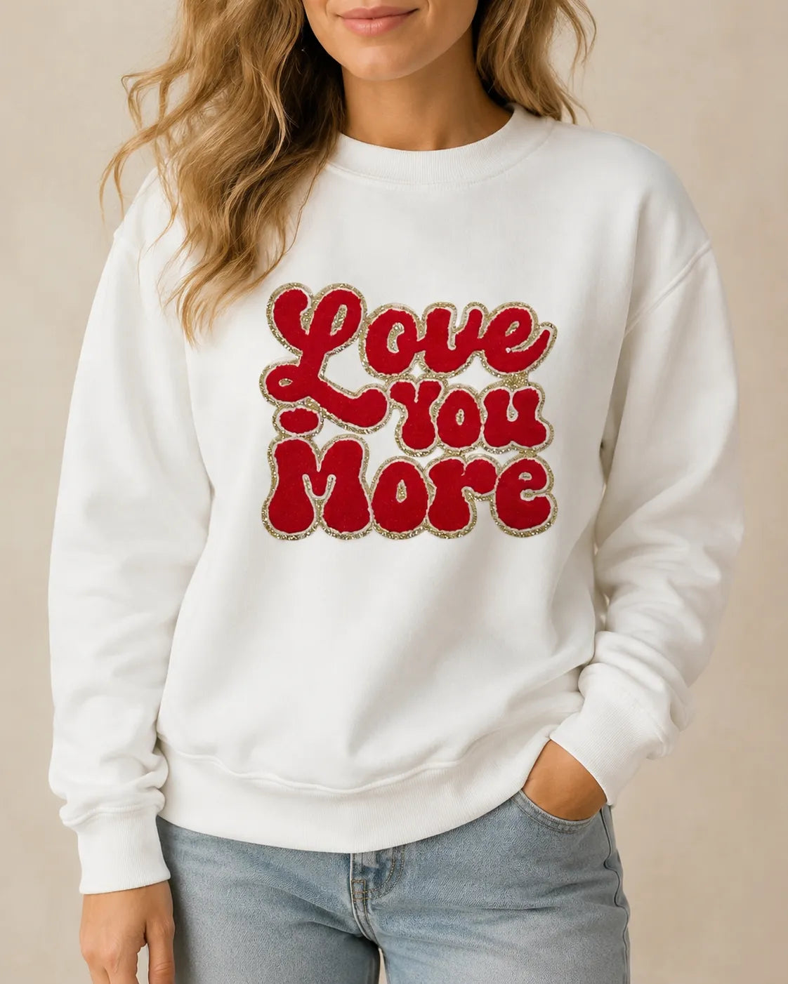 A woman wearing a white crewneck sweatshirt with 'Love You More' printed in red retro-style lettering with a glittery outline, paired with light blue jeans against a neutral beige background.